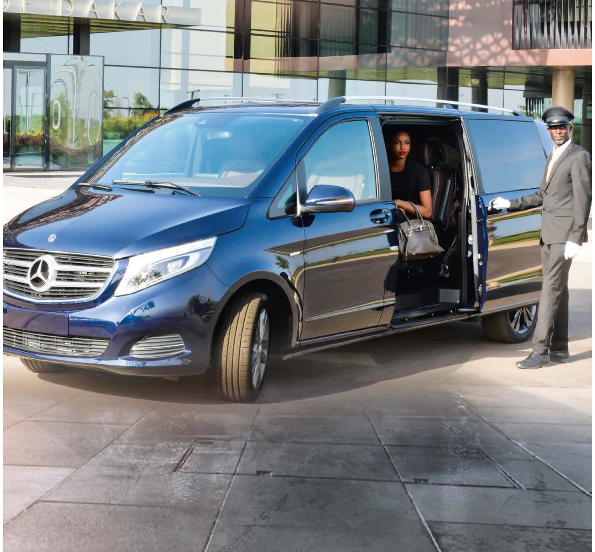 Chauffeur ouvrant la porte coulissante d'un van Mercedes bleu nuit devant un hôtel, une passagère à bord